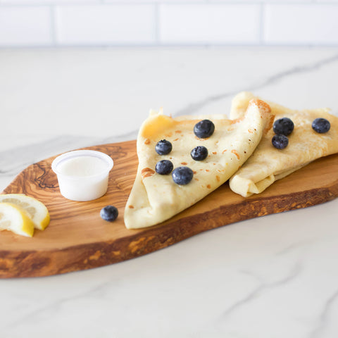 Two crepes with blueberries on a wooden board with lemon slices and a small container of cream.