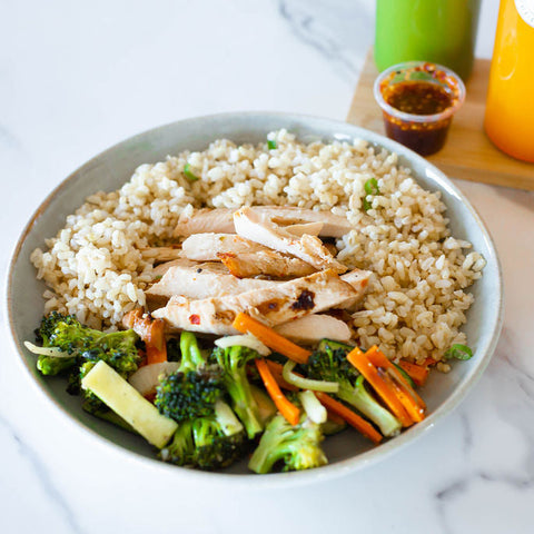 Bowl of quinoa salad with chicken, broccoli, and carrots on a marble surface with drinks in the background.